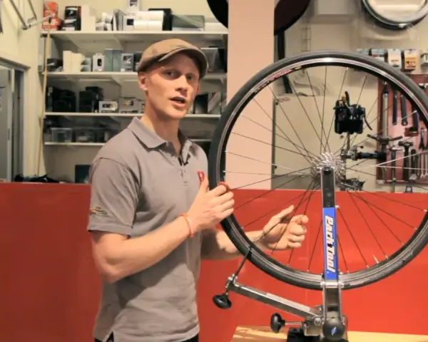Man i keps visar hur man rikta cykelhjul i en verkstad. Verktyg hänger på väggen bakom.