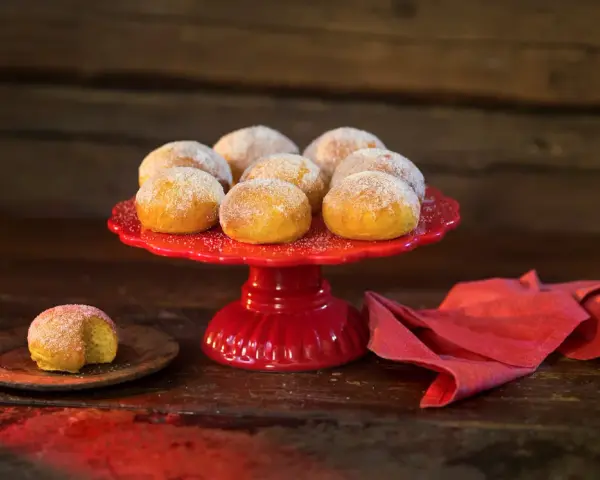 En röd tårtfat med lussebullar med kvarg, pudrade med socker. En ensam bulle ligger på en träplatta.
