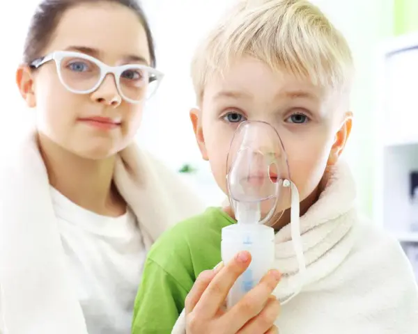 Dziecko z maską inhalatora, obok dziewczynka w okularach.
