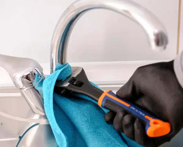 Mano enguantada usa llave inglesa para cambiar grifo cocina. Toalla azul protege el cromo.
