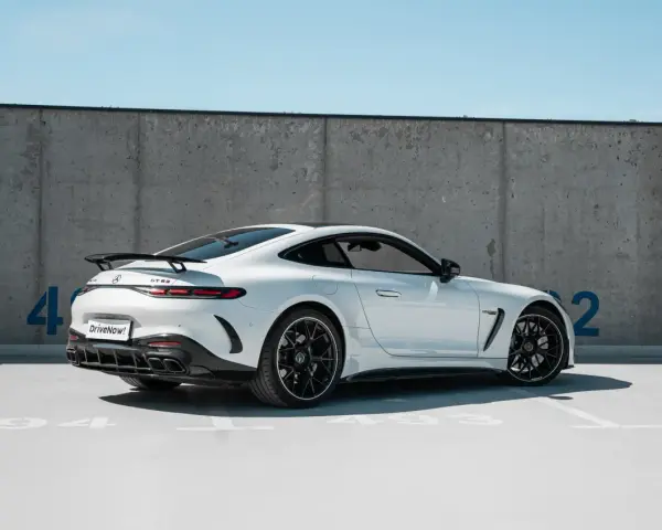 Biały Mercedes GT 63 z czarnym spojlerem i felgami, zaparkowany pod betonową ścianą z niebieskimi cyframi.