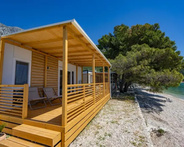 Domek mobilny z drewnianym tarasem i widokiem na morze w camp Dole Živogošće. Dwa leżaki czekają na plaży.