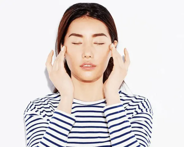 A woman with closed eyes performs a gentle face massage for puffiness, her fingers delicately touching her temples.