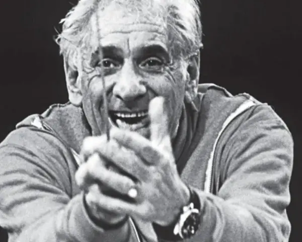 Uśmiechnięty Leonard Bernstein, dyrygent, z dłońmi złożonymi przed sobą, wpatruje się w widza.