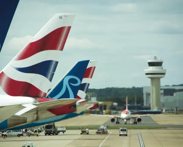Samoloty z charakterystycznymi ogonami British Airways i innym logo czekają na start na lotnisku Londyn Gatwick. W tle wieża kontroli lotów.