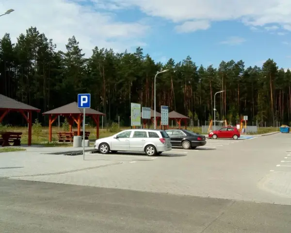 Parking przy lesie, z wiatami i ławkami. W tle las, a na parkingu kilka samochodów. To idealne miejsce na postój w drodze, może nawet w pobliżu **Orlen Sople**.