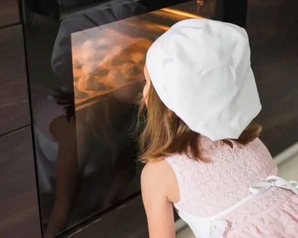 Niña con gorro de chef mirando dentro de un horno encendido.