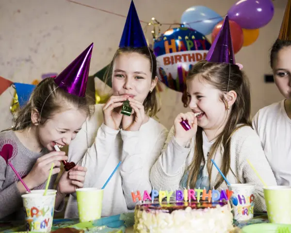 Radosne zabawy na urodziny! Dziewczynki w stożkowych czapkach dmuchają w gwizdki, śmiejąc się przy torcie z napisem "Happy Birthday".