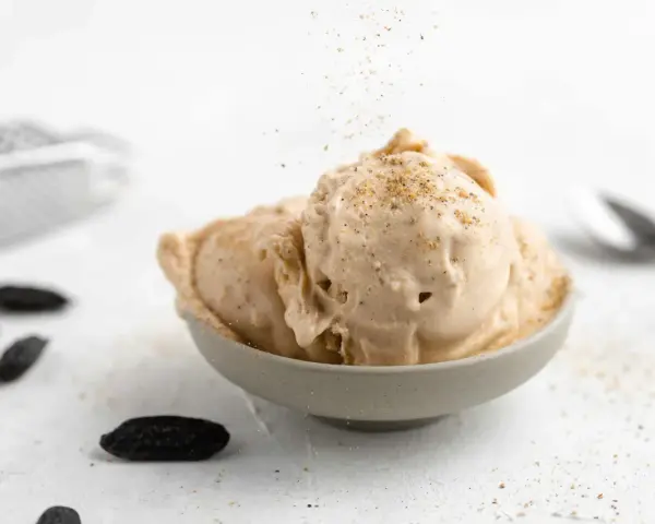 Cremiges Tonkabohnen Eis in einer Schale, bestreut mit gemahlenen Tonkabohnen. Mehrere ganze Tonkabohnen liegen daneben.
