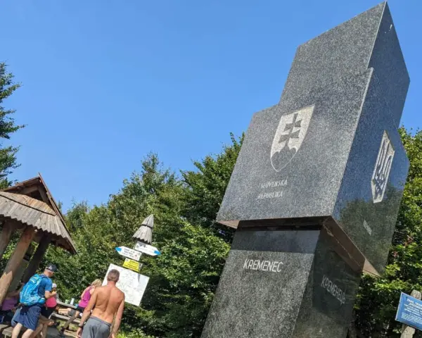 Trójstyk granic Bieszczady: monumentalny obelisk z herbami Słowacji i Ukrainy, obok drewniana wiata i turyści.