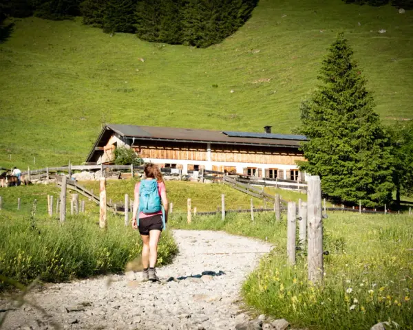 Frau mit Rucksack wandert auf einem Schotterweg zu einer Berghütte. Grüne Wiesen und Bäume im Hintergrund.