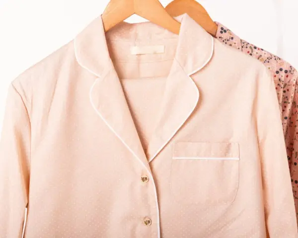 Pyjama rose à pois blancs, col rabattu, suspendu sur un cintre en bois. Une autre chemise à fleurs est visible derrière.