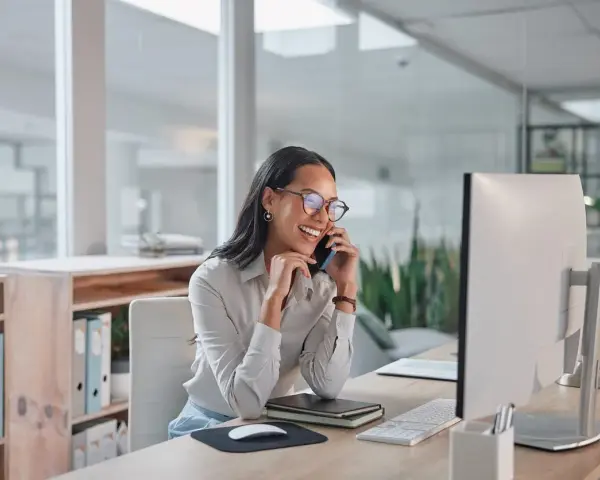 Donna sorridente al telefono in ufficio, mentre lavora al computer. Potrebbe chiedersi quanto guadagna una segretaria al mese.