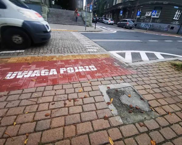 Czerwony pas na chodniku z napisem "UWAGA POJAZDY" informuje o przejściu sugerowanym. W tle widać samochody i schody.