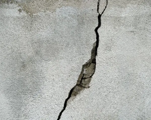 Pękające ściany w nowym domu, widoczna głęboka szczelina w jasnoszarym tynku, odsłaniająca fragmenty betonu.