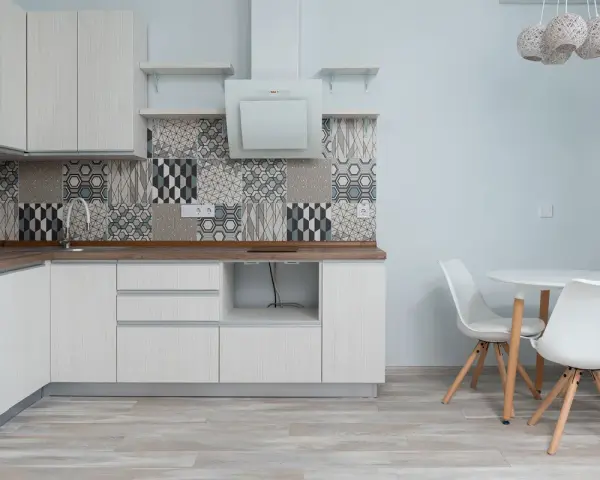 Cocina moderna con armarios blancos, salpicadero geométrico y suelo gris claro. Mesa redonda con sillas blancas.