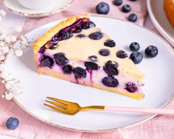 Ein Stück Heidelbeerkuchen Landfrauenart mit saftigen Blaubeeren und cremigem Guss auf einem Teller, daneben eine Gabel und frische Beeren.