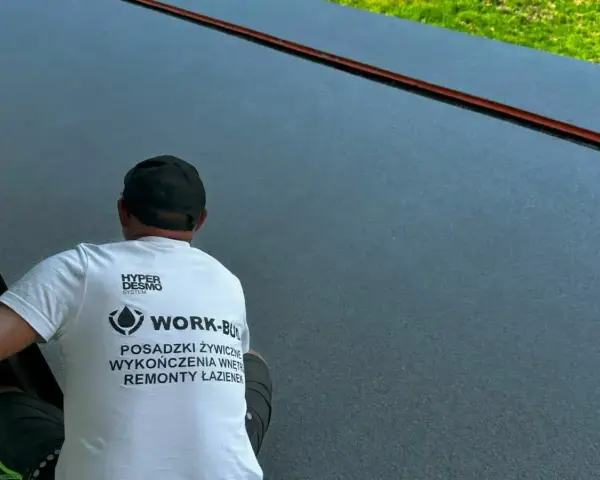 Pracownik w koszulce z napisem "Posadzki żywiczne" wykonuje prace na ciemnej, gładkiej powierzchni.