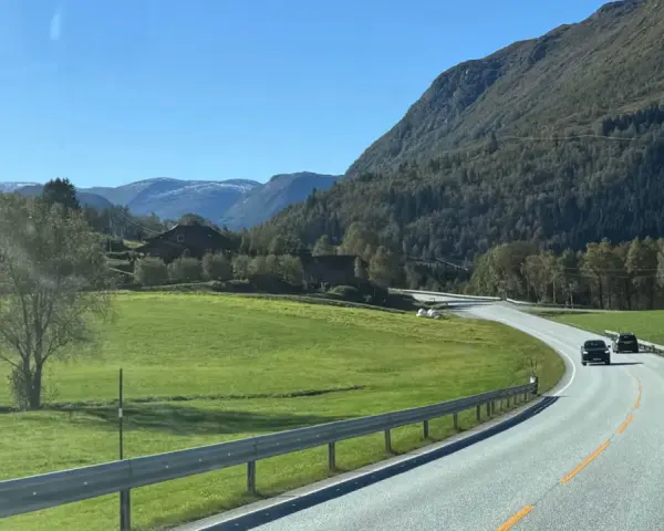 En bil kör på en slingrande väg genom ett grönt landskap med berg och hus. En perfekt start på en semester i Norge.