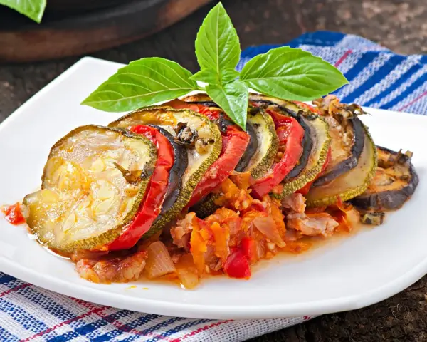 Ratatouille con rodajas de calabacín, berenjena y tomate, decorado con albahaca. Los ingredientes ratatouille se ven tiernos y jugosos.