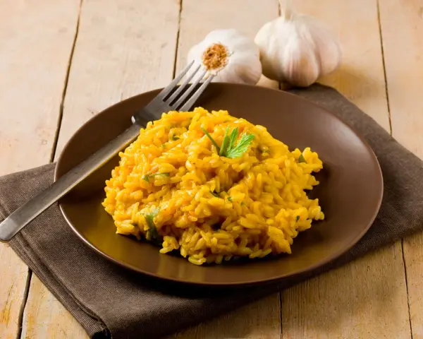 Un invitante risotto zafferano in polvere, guarnito con prezzemolo fresco, su un piatto marrone, con aglio e forchetta sullo sfondo.