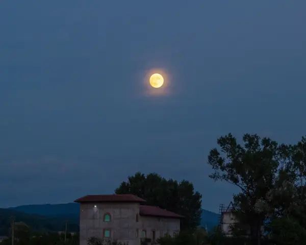 La luna di luglio illumina un paesaggio serale con colline, alberi e un edificio.