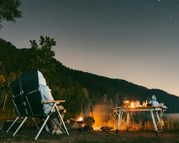 Personne assise près d'un feu de camp sous un ciel étoilé, avec une table de camping et un lac.
