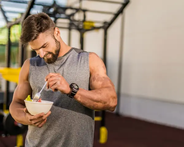 Hombre con barba en gimnasio, comiendo ensalada. Demuestra que se puede ganar músculo en déficit calórico con la dieta adecuada.