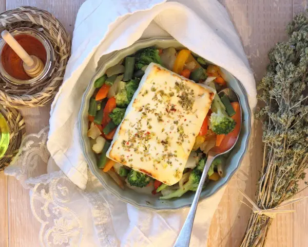 Feta al forno con verdure miste, condita con erbe aromatiche. Accanto, olio, spezie e un mazzetto di origano secco.