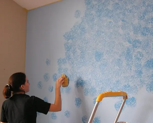 Mujer aplica pintura que disimula imperfecciones en pared con esponja, creando un patrón de nubes azules.