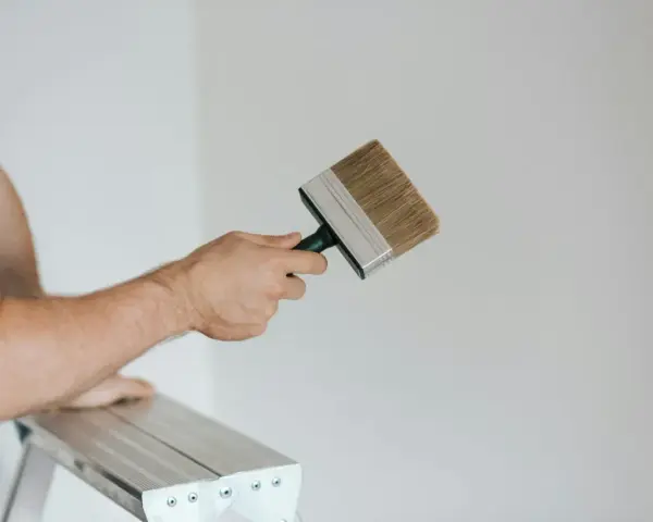 Person på en stege håller en bred pensel redo att måla huset vitt.