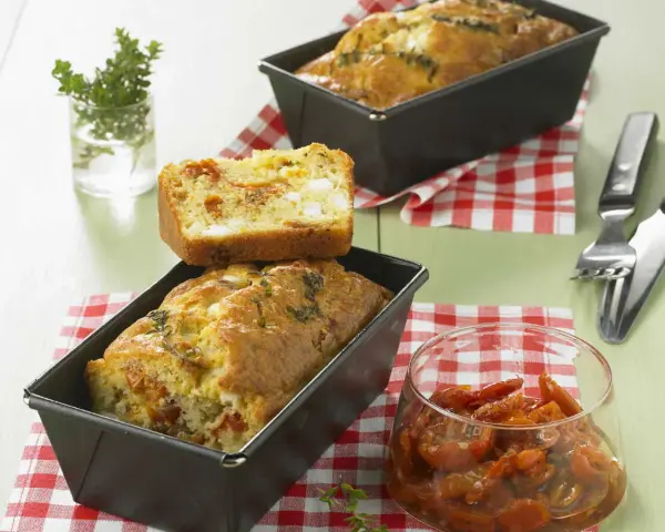 Cake tomates séchées feta, un délice salé aux herbes fraîches. Une tranche est posée sur un cake encore dans son moule.