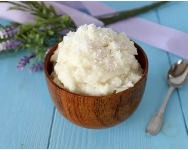 Una ciotola di legno piena di cremosa crema al cocco, guarnita con cocco grattugiato. Accanto, un cucchiaino d'argento e fiori di lavanda.