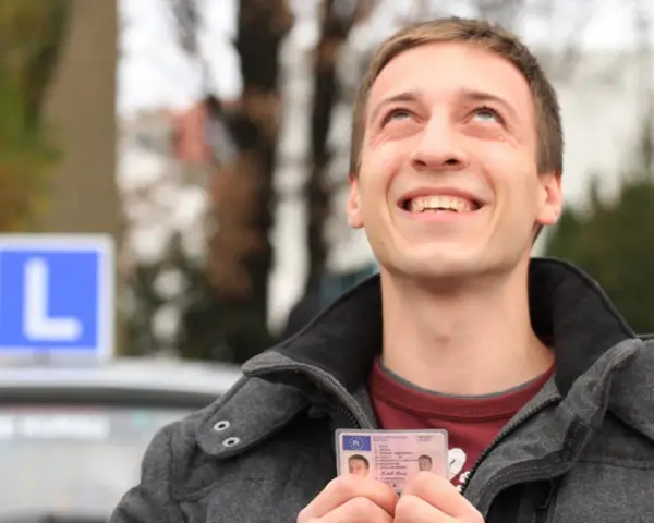 Szczęśliwy młody mężczyzna trzyma prawo jazdy kategorii B, z niebieskim znakiem "L" na dachu samochodu w tle.