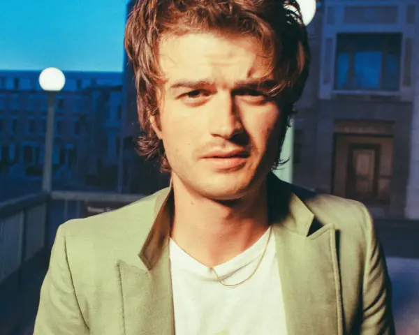 Joe Keery w jasnej marynarce, z naszyjnikiem, na tle nocnego miasta.