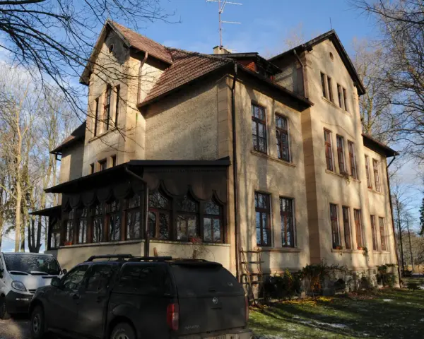 Dawny pałac Poraj, z charakterystyczną werandą i drzewami w tle, otoczony zimowym krajobrazem.