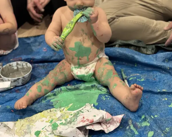 Bébé couvert de peinture verte, tenant un pinceau, assis sur une bâche bleue.
