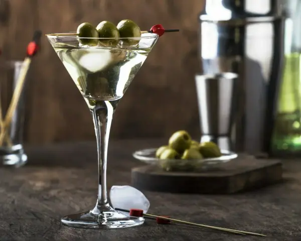 Elegancki martini drink z trzema oliwkami na patyczku, lodem i akcesoriami barmańskimi w tle.