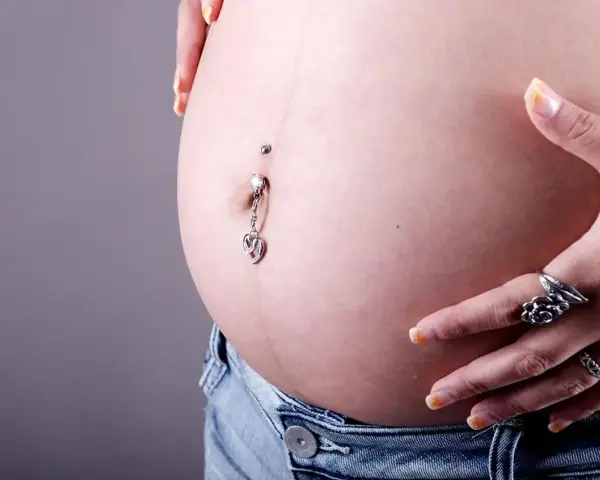 Brzuszek w ciąży z ozdobnym kolczykiem w pępku. Warto pamiętać, że kolczyk w pępku w ciąży może być problematyczny.