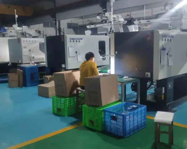 A person in a yellow jacket works at a station with boxes and crates, surrounded by injection molding machines, indicating low volume production.