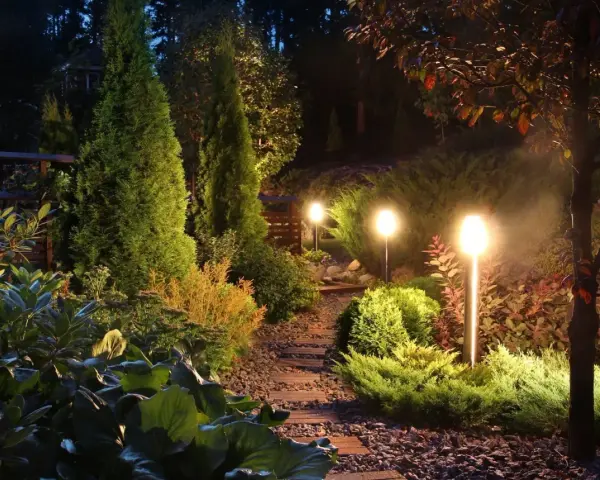 Oświetlenie bez prądu tworzy magiczną atmosferę w ogrodzie. Ścieżka z kamieni i drewna prowadzi przez bujną roślinność, oświetloną ciepłym blaskiem lamp.