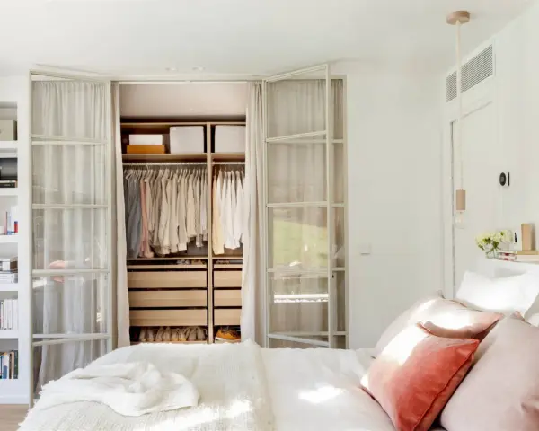 Dormitorio con vestidor integrado. Ropa organizada en armarios abiertos y estanterías. Cama con cojines rosas y manta blanca.