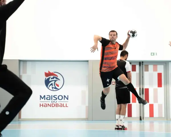 Piłkarz ręczny, niczym szczypiornista, wyskakuje do rzutu. W tle logo "Maison du Handball".