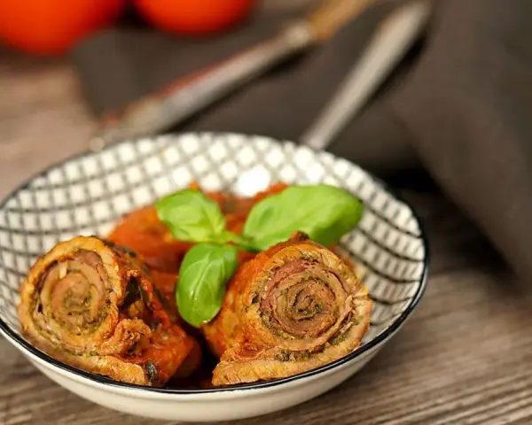 Zwei herzhafte involtini originalrezept in Tomatensoße, garniert mit Basilikum, in einer Schale auf Holztisch.