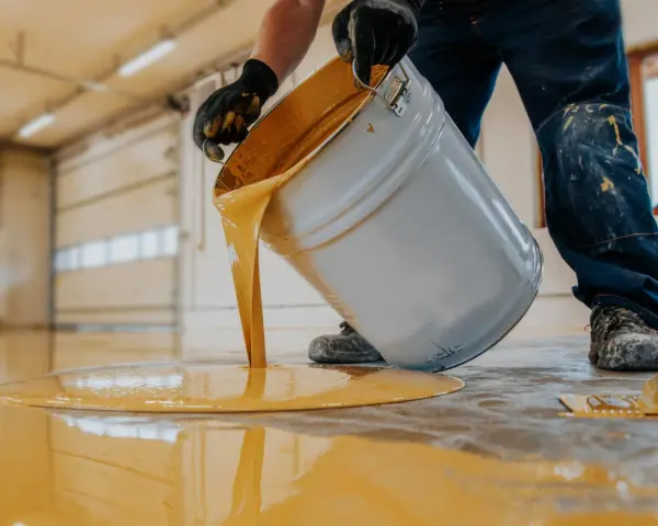 Hombre con guantes vierte pintura para suelo de cemento de color amarillo brillante de un cubo.