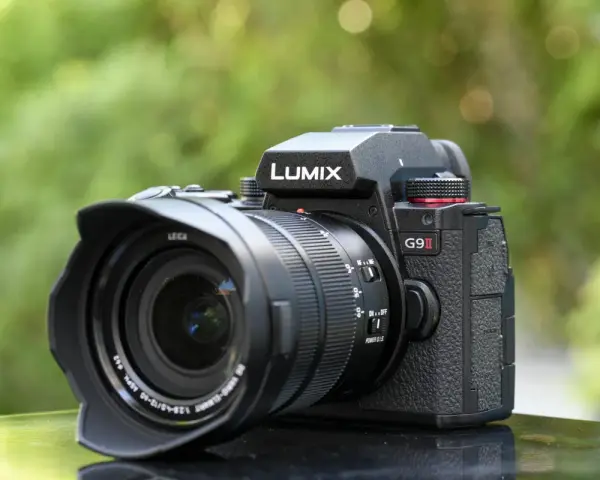 Un appareil photo noir Lumix G9 II avec objectif Leica est posé sur une surface réfléchissante, avec un arrière-plan flou de verdure.