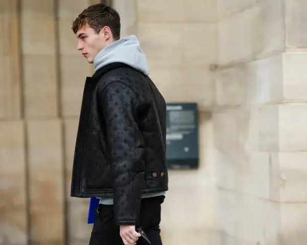 Uomo con un elegante outfit con giubbotto di pelle nero con monogrammi, felpa grigia e pantaloni scuri.