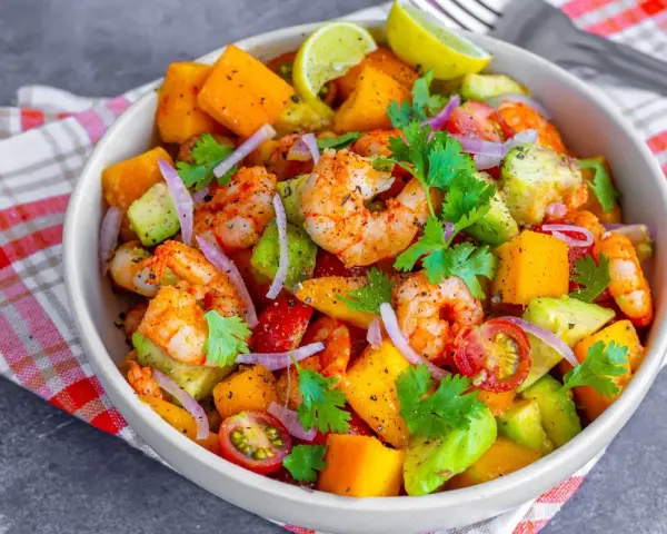 Kolorowa sałatka z krewetkami, awokado, mango, pomidorkami i czerwoną cebulą, udekorowana kolendrą i ćwiartkami limonki.