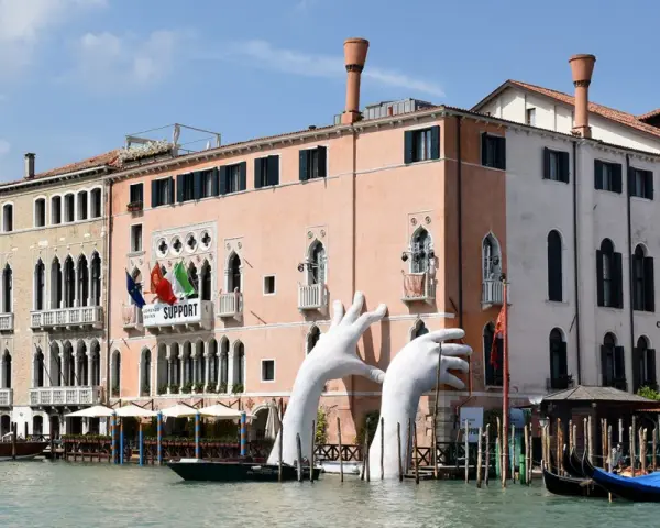 Enorme installazione di mani bianche emerge dal Canal Grande, davanti al museo arte contemporanea Venezia. Gondole ormeggiate.