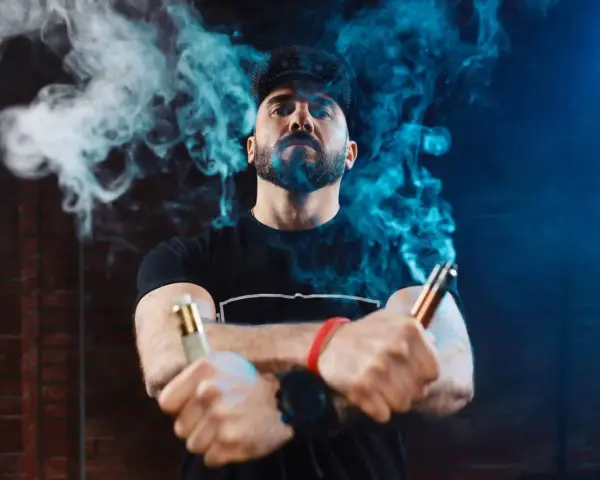 Hombre con barba y gorra, rodeado de vapor azul, sostiene dos vapers, uno dorado y otro cobre. La diferencia entre 0.8 y 1.2 ohm se siente en la calada.